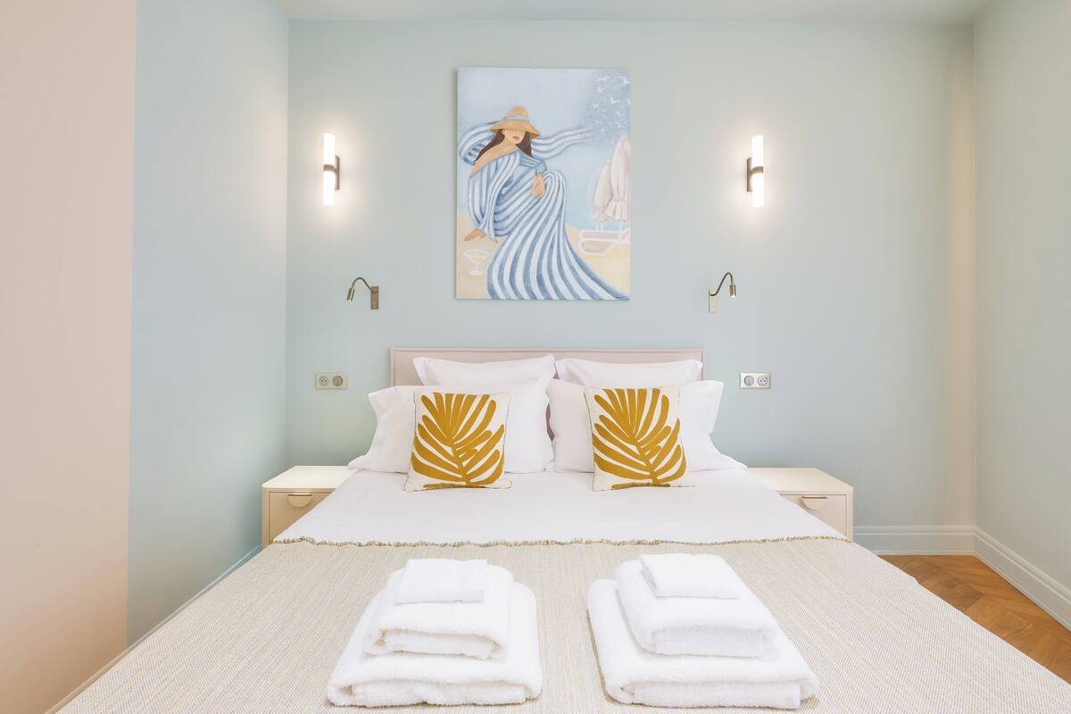 A neatly made bed with white linens and two pillows, a painting above the bed, and two wall-mounted lights.