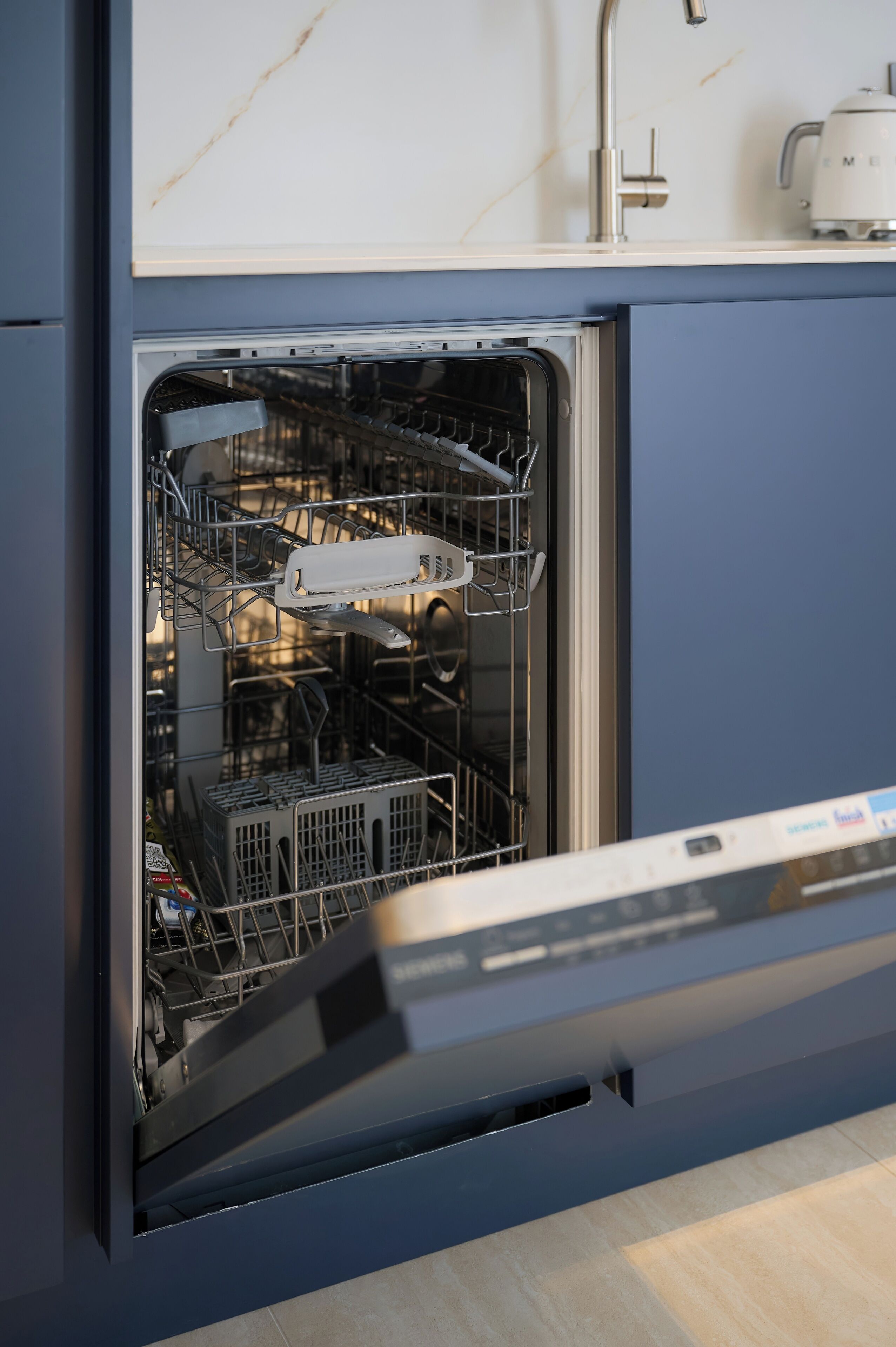 An open dishwasher with racks and utensils inside, a white coffee maker on the counter, and a marble backsplash.