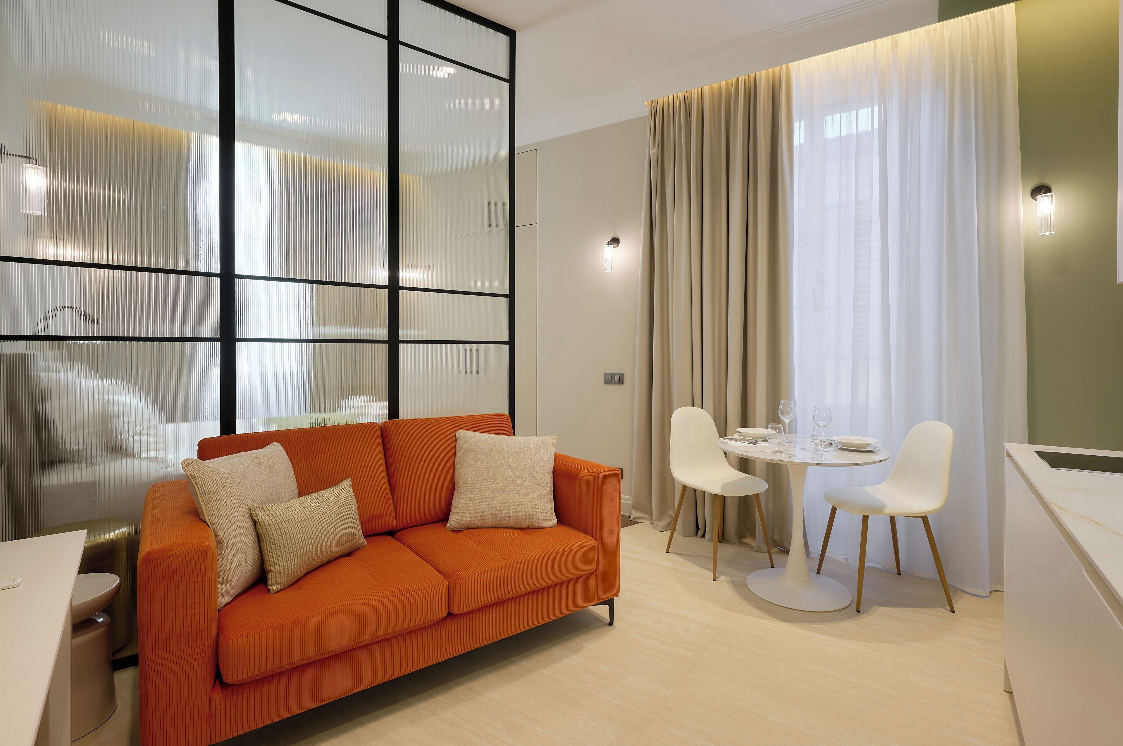 A modern hotel room with a sofa, dining table, chairs, and a large window with curtains.