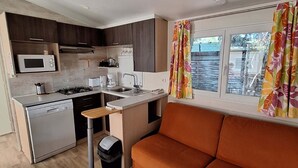 Cocina compacta con microondas, estufa y lavabo, un pequeño cubo de basura y un sofá cerca de una ventana con cortinas.