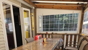 Un área de patio techada con una mesa larga y sillas, piso de madera y ventanas amplias que ofrecen vistas al exterior.