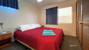 A bedroom with a bed, a nightstand, a lamp, a ceiling fan, and a window with blinds. - Burrum River Unit One (Burrum Heads)