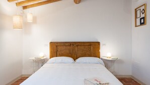 Un dormitorio con cabecera de madera, ropa de cama blanca, dos mesitas de noche con lámparas y una pequeña mesa redonda con un libro.