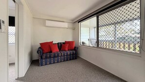 Une pièce avec un canapé bleu à motifs, des coussins rouges, une grande fenêtre avec un grillage métallique et un climatiseur blanc fixé au mur.