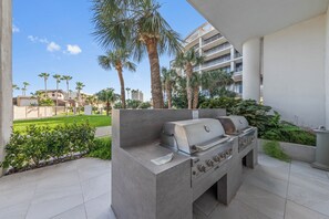 An outdoor kitchen area with a built-in grill, surrounded by palm trees and greenery, with a view of a modern building in the background. - Spacious, 2-Story Condo in Beachfront Resort – Pools, Hot Tubs, Grills, & More! (South Padre Island)