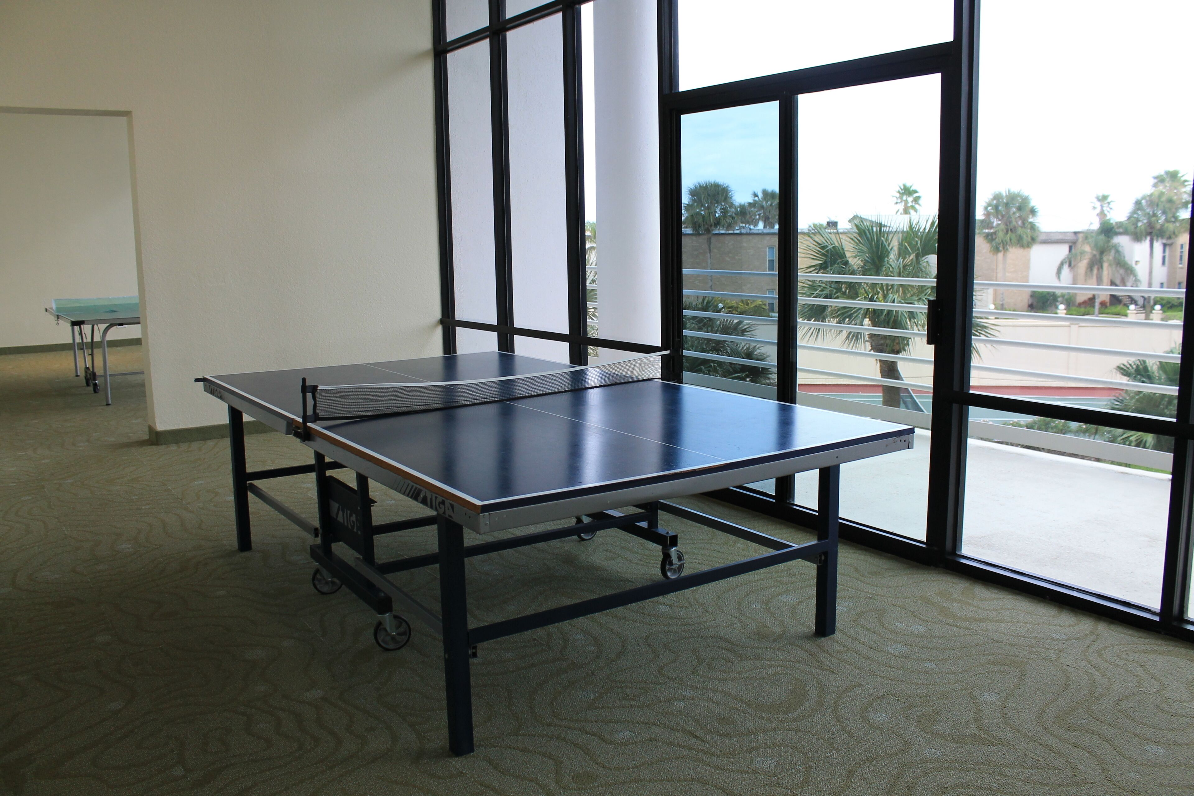 Una mesa de ping pong en una habitación con ventanas amplias y vistas a palmeras afuera.