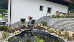 En damm med en kant av stenar, omgiven av småsten och växter, med ett hus i bakgrunden.