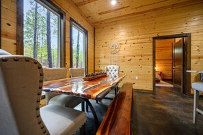 Interior de una cabaña de madera con una mesa de comedor, sillas y vistas al bosque a través de amplios ventanales.