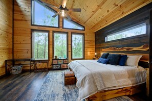 A wooden cabin bedroom with a bed, large windows, a ceiling fan, and a rustic wall design. - Moonshine Times - great outdoor space, hot tub, playground, firepit (Broken Bow)