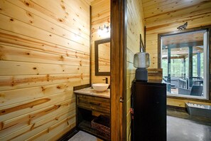 Ett badrum i en stuga med träpanel på väggarna, ett vitt handfat, en spegel och ett fönster med utsikt över naturen.