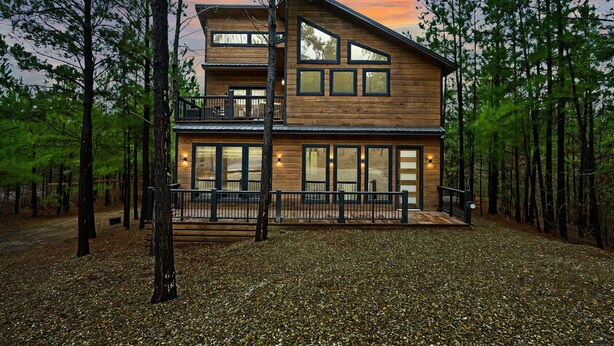 A two-story wooden cabin with a balcony, surrounded by trees. - Moonshine Times - great outdoor space, hot tub, playground, firepit (Broken Bow)