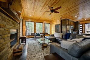 A cozy cabin interior with a stone fireplace, leather sofa, and a large window overlooking a forest.