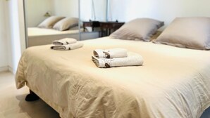 Una cama bien hecha con una colcha color beige y dos almohadas, una mesita con una silla y un espejo en la pared.