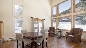 A spacious room with a wooden dining table surrounded by chairs, large windows with a snowy view, and a fireplace. - Spacious Home Base! Hot Tub, Fire Pit, Sleeps 10 (Avon)