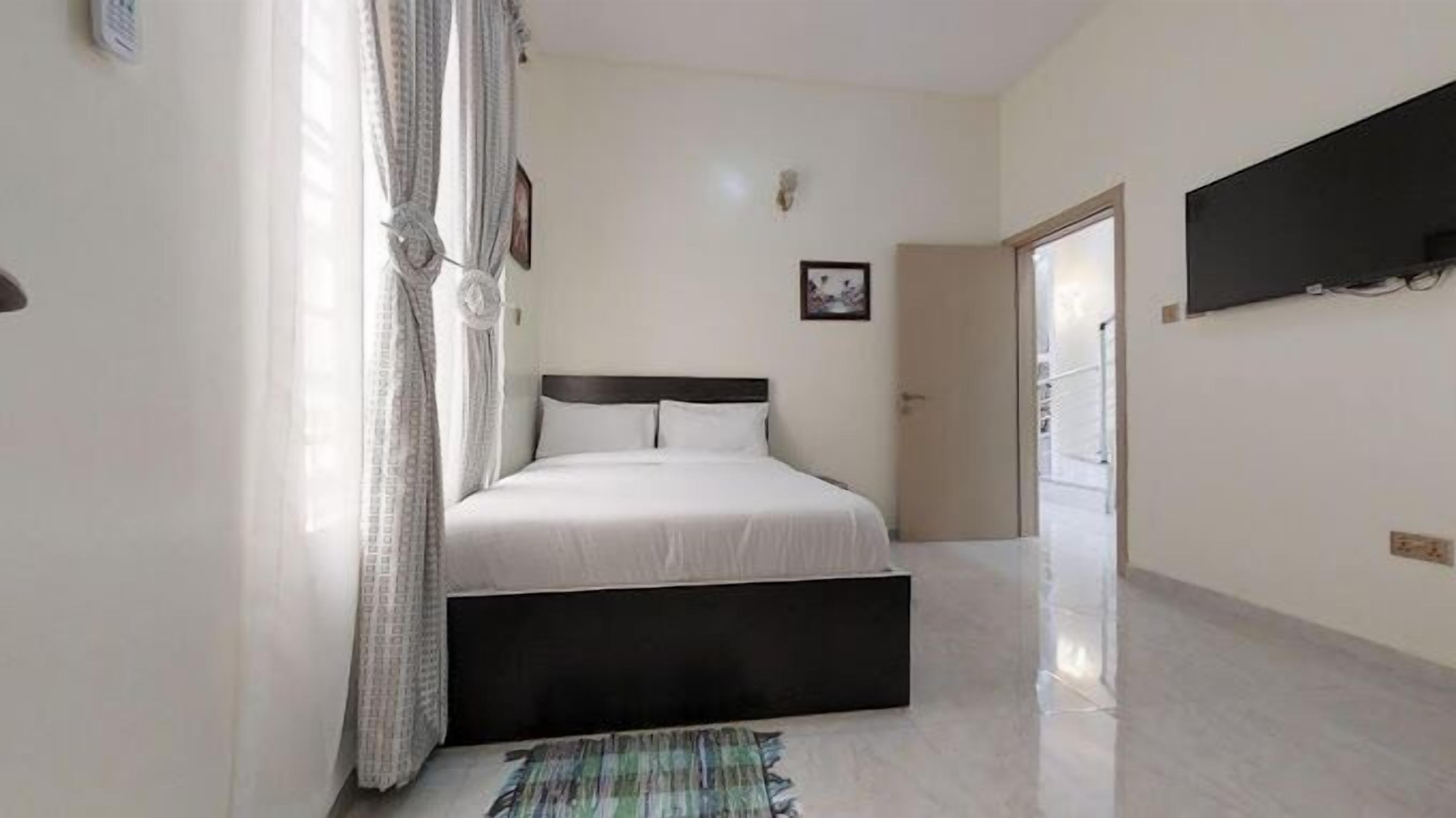 A bedroom with a bed, curtains, a flat-screen TV, and a door leading to another room.