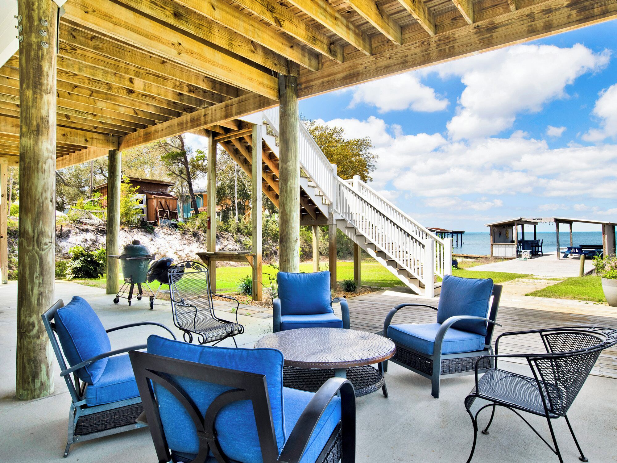 A covered outdoor patio with blue chairs and a round table, a wooden staircase, and a view of the ocean.