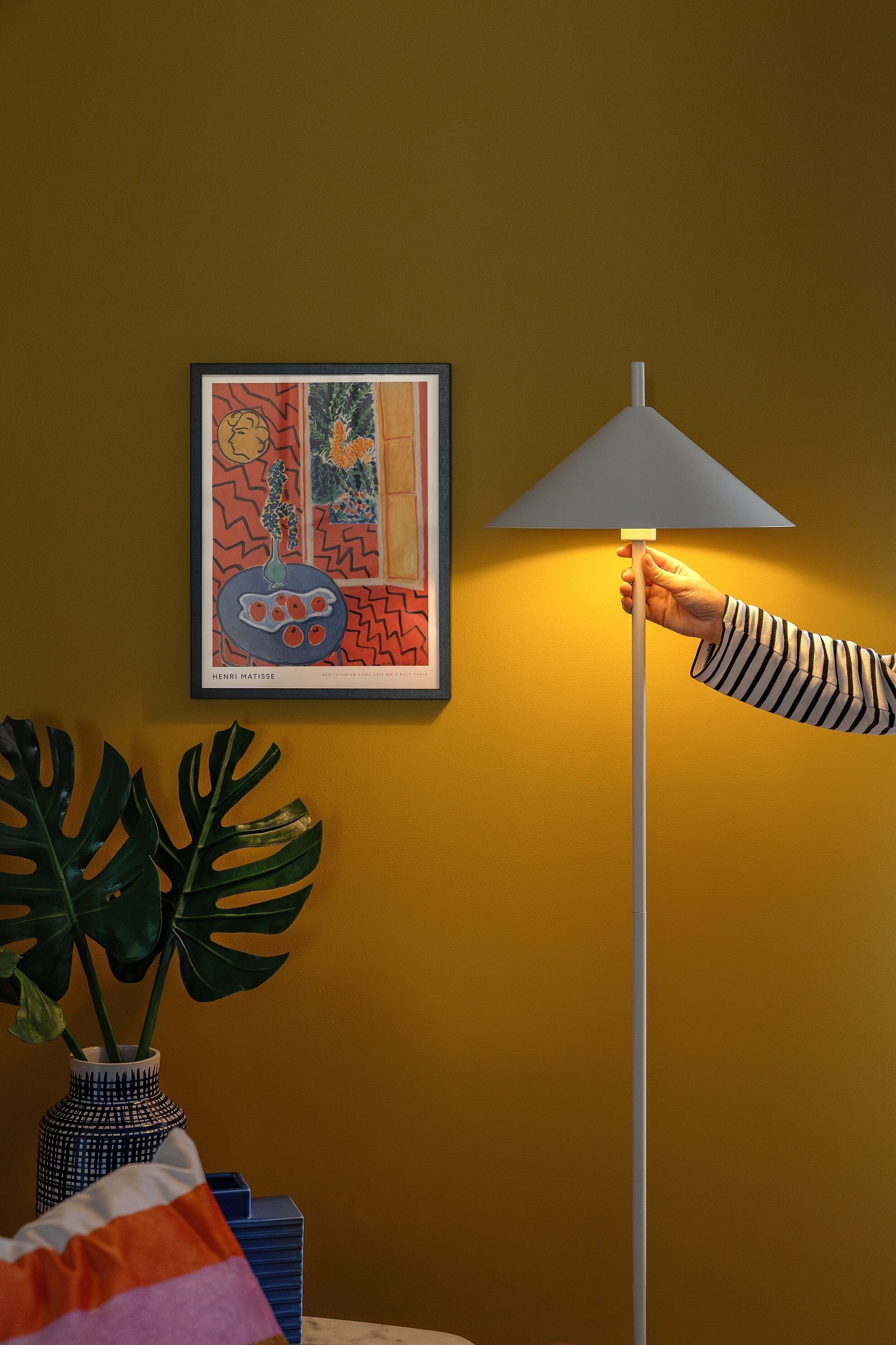 Une pièce avec un tableau encadré au mur, une plante en pot et une lampe.