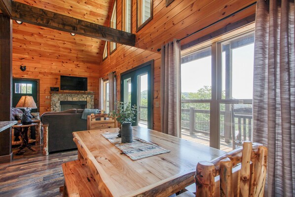 A wooden cabin interior with a dining area, a fireplace, and large windows.