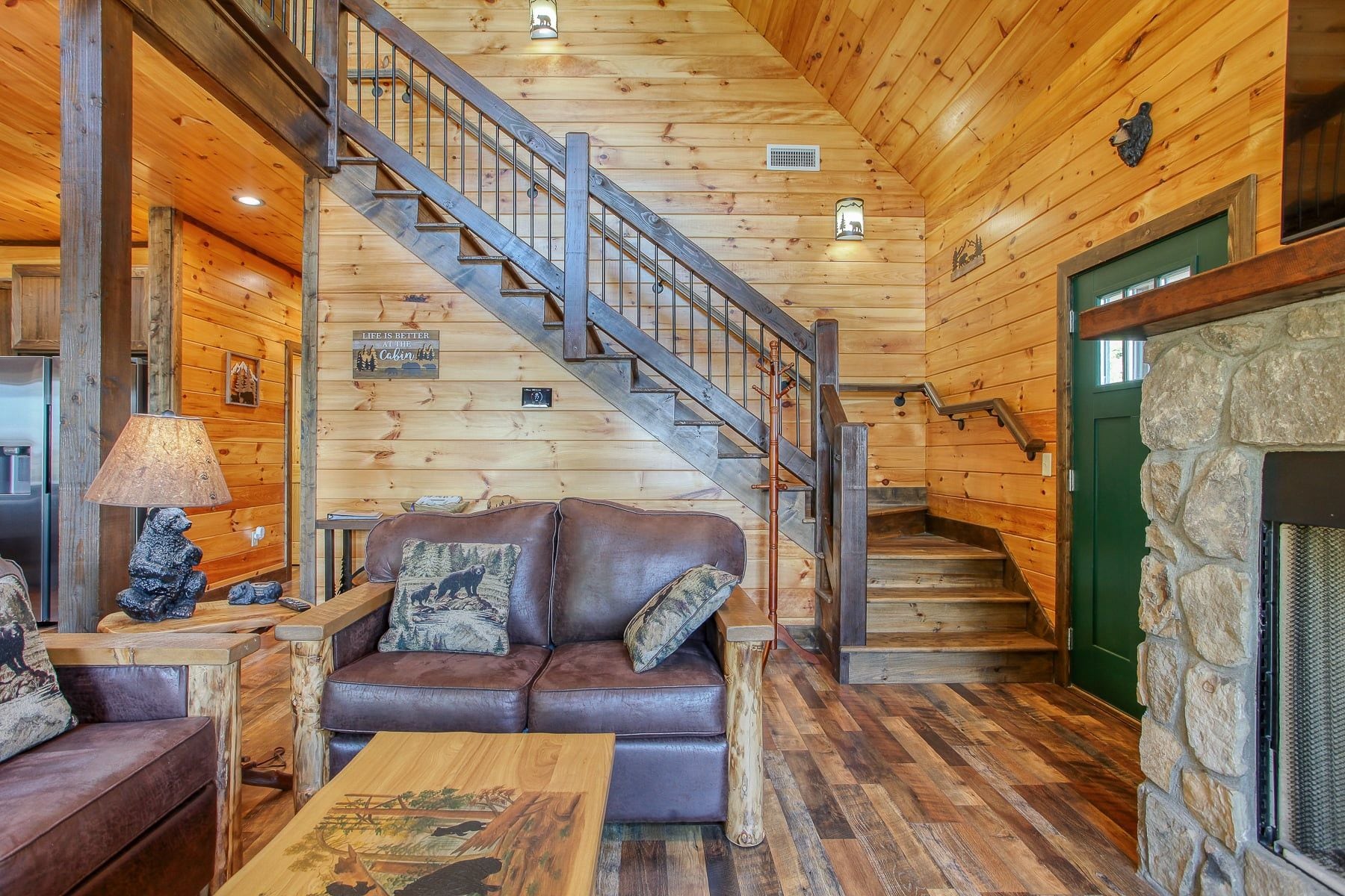A cabin interior with a wooden staircase, leather sofa, and a stone fireplace.