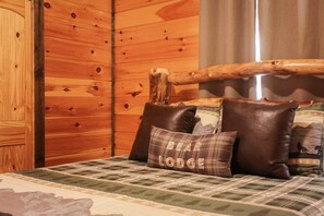 A wooden cabin interior with a bed featuring a plaid bedspread and several pillows, one with 'BEAR LODGE' text.