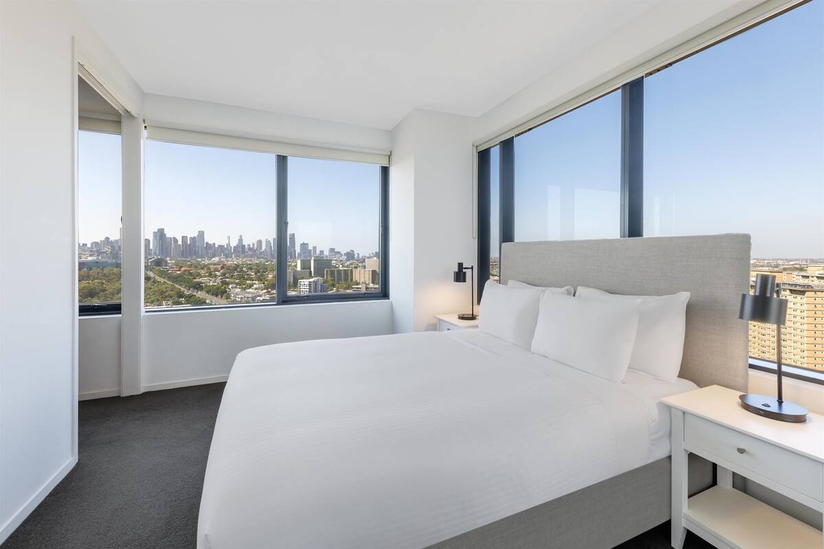 A modern hotel room with a large bed, a nightstand, and a view of a city skyline through large windows.