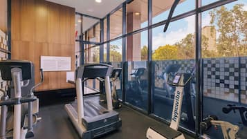 Un gimnasio con cintas de correr, elípticas y máquinas de pesas, amplias ventanas con vistas a árboles y edificios, y una pared de madera con carteles informativos.