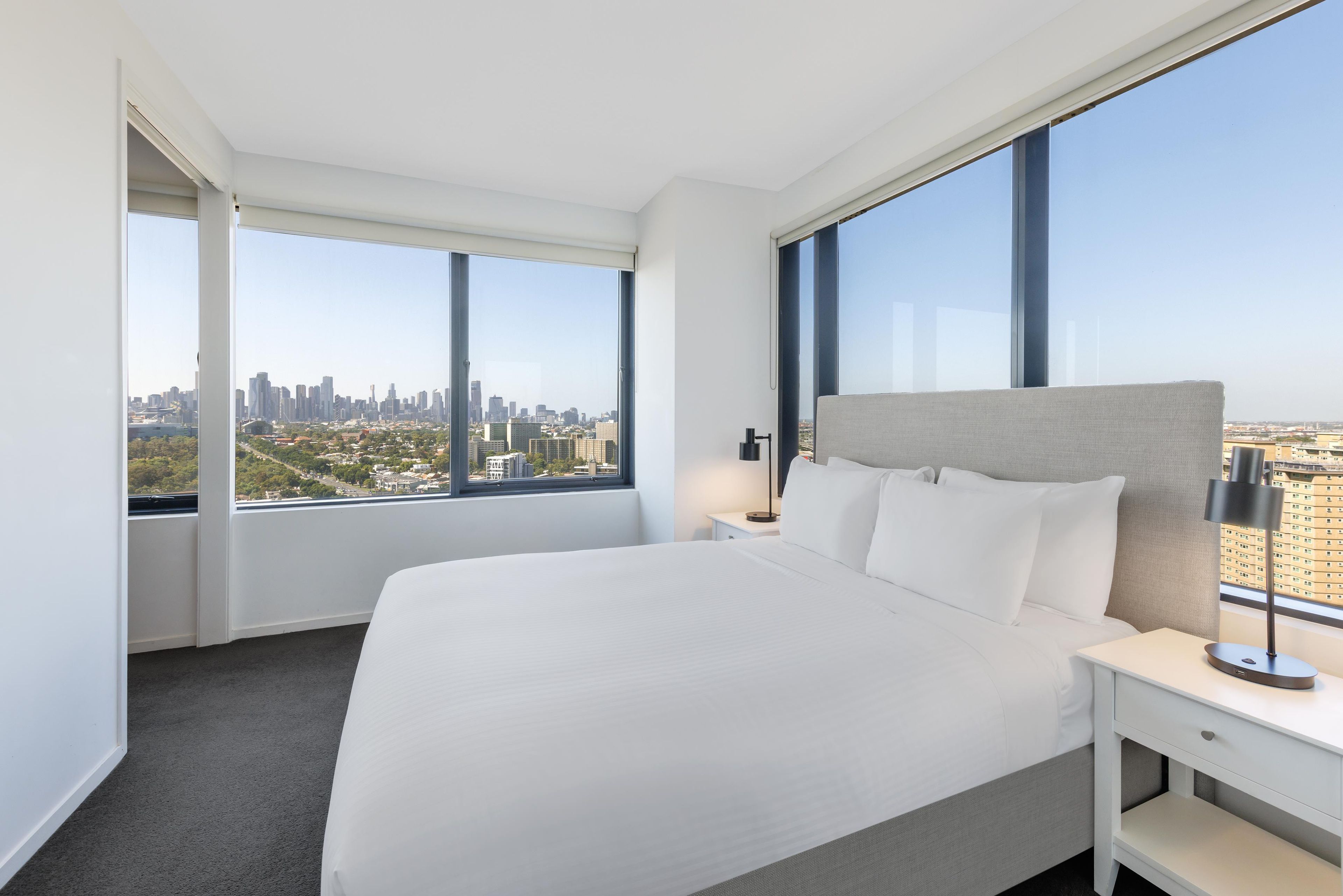 Habitación de hotel con una cama grande, una mesita de noche, una lámpara y vistas a un paisaje urbano a través de amplias ventanas.