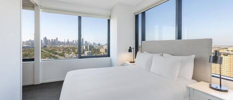 A hotel room with a large bed, a nightstand, a lamp, and a view of a cityscape through large windows.