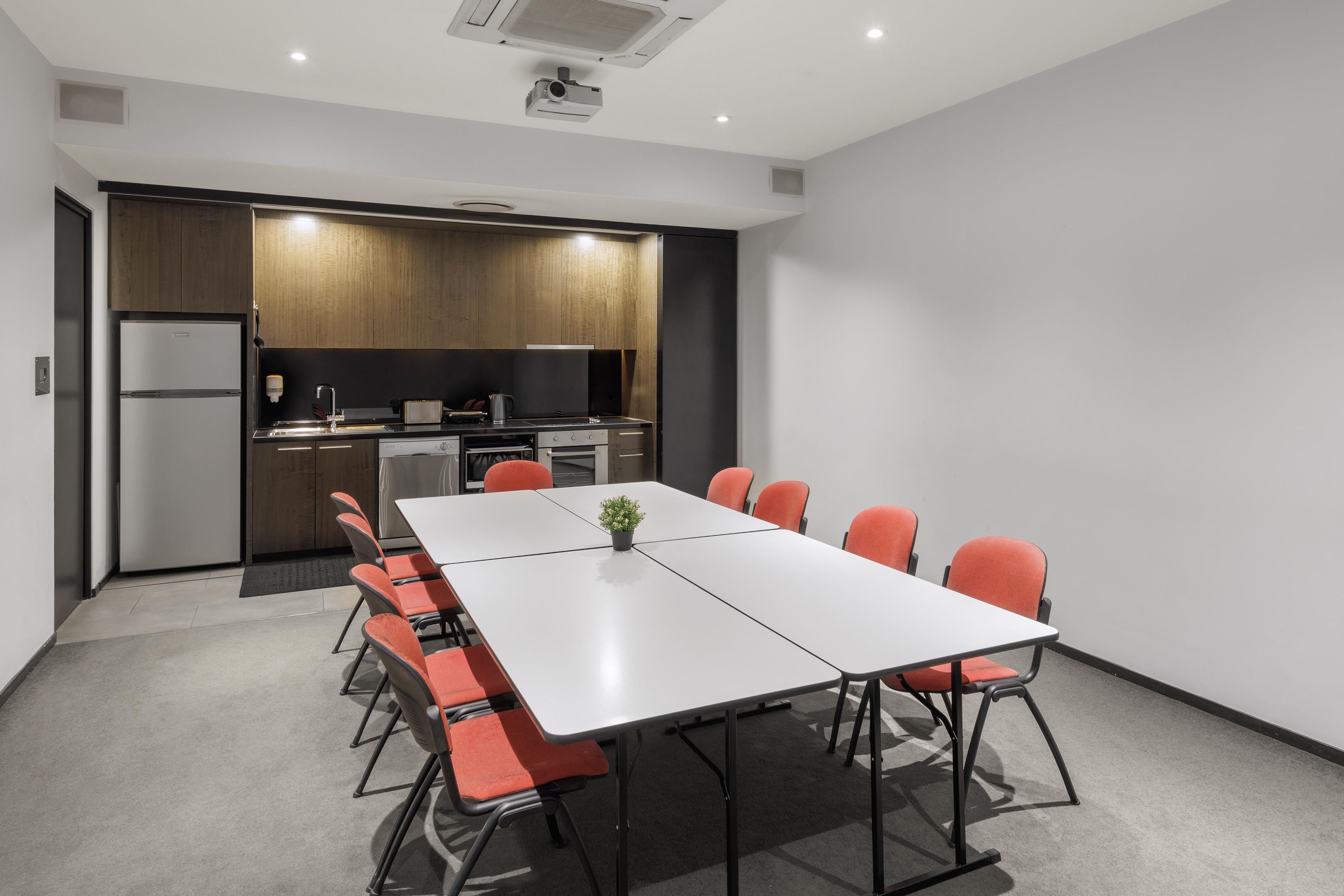 A modern meeting room with a long table and orange chairs, a kitchen area with wooden cabinets, and a built-in projector.