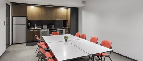Una sala de reuniones moderna con una larga mesa y sillas anaranjadas, una zona de cocina con armarios de madera y un proyector integrado.