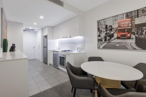A modern kitchen with white cabinets, a built-in oven, and a dining area with a round table and chairs. - Oaks Melbourne Flemington Suites (Travancore)