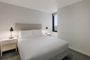 Habitación de hotel minimalista con una cama grande, dos mesitas de noche, una mesita auxiliar, un ventanal con vista a la ciudad y suelo alfombrado.