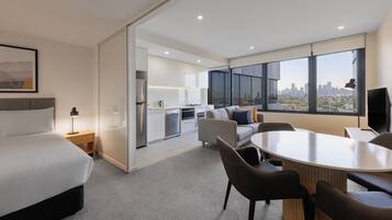 A modern hotel room with a bed, a desk, a dining table, and a sofa. There is a kitchen area with a refrigerator, oven, and microwave. The room has large windows with a city view.