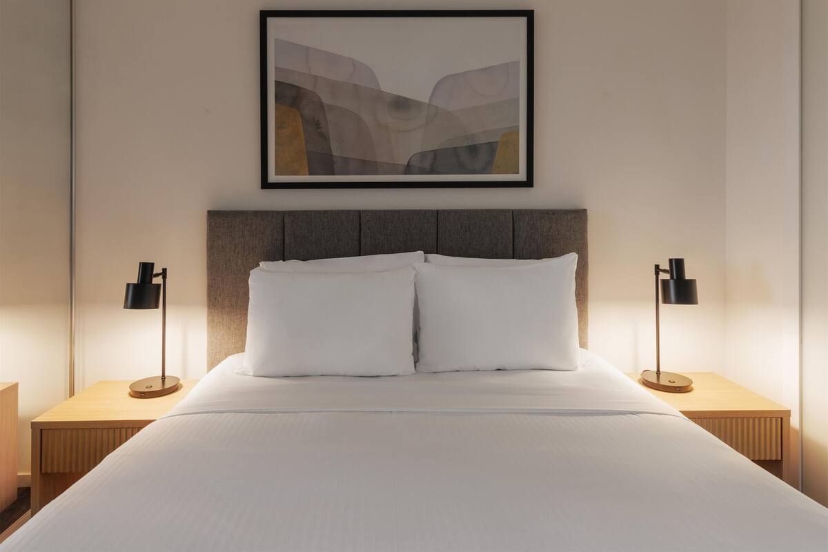 A neatly made bed with white linens, two bedside lamps, and a framed abstract painting on the wall.