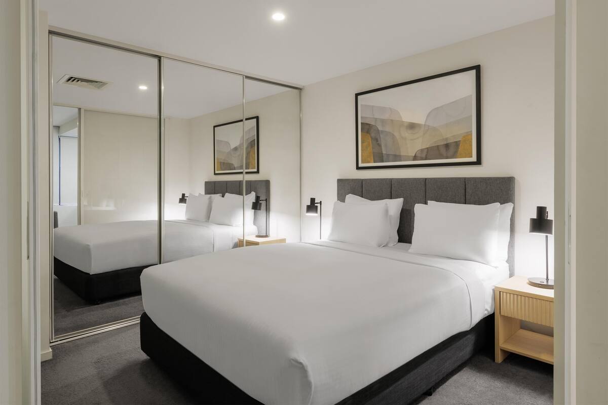A modern hotel room with a large bed, a bedside table with a lamp, a mirror, and a framed abstract painting on the wall.
