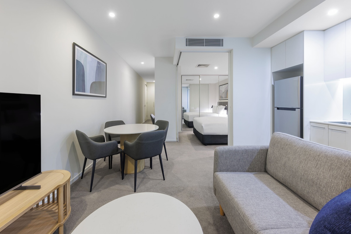 Una habitación de hotel moderna con una televisión de pantalla plana, una mesa de comedor redonda con sillas, un sofá gris y una cama con colchón blanco.