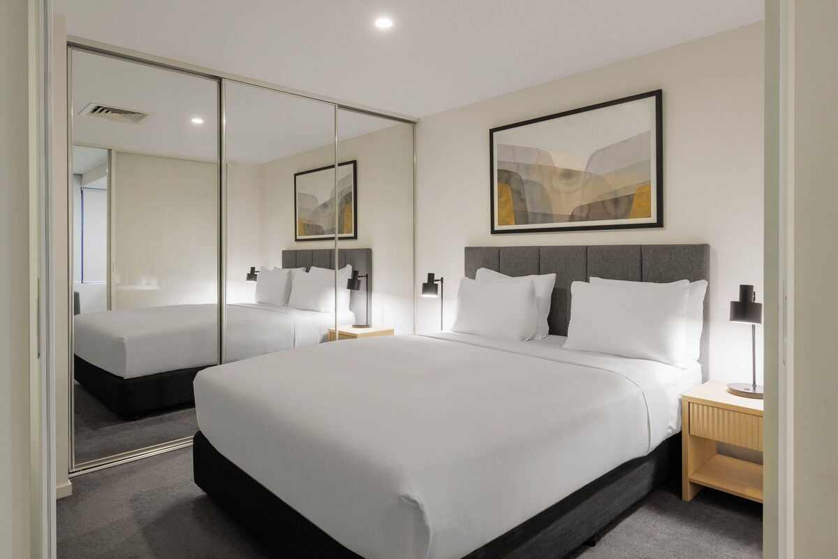 Une chambre d’hôtel moderne avec un grand lit, une table de chevet, une commode, un miroir et un tableau abstrait encadré accroché au mur.