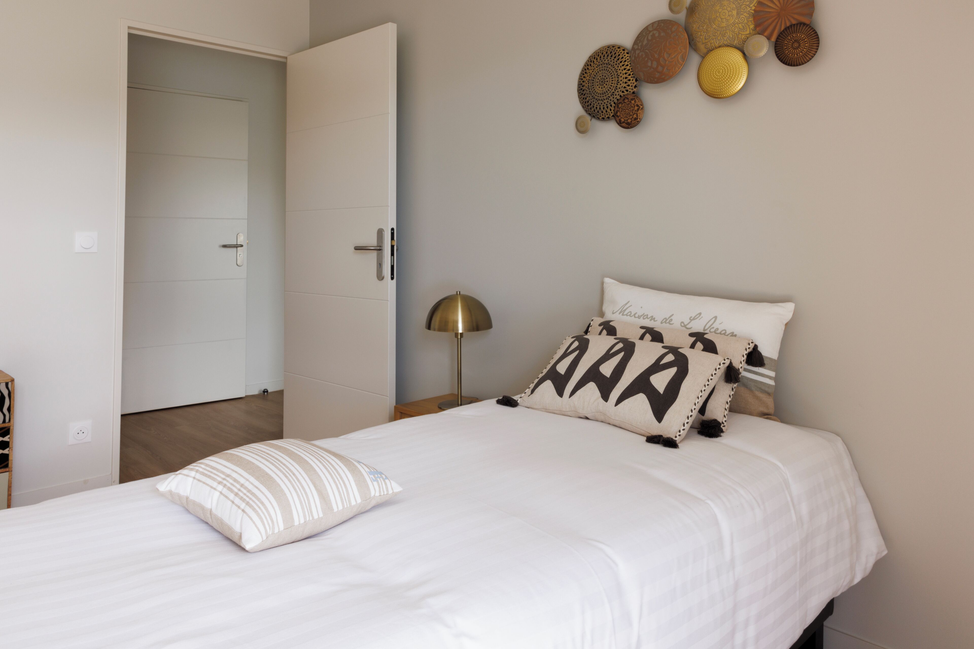 A neatly made bed with a white comforter and two pillows, one with a geometric design and another with text. A bedside lamp is on the nightstand beside the bed.
