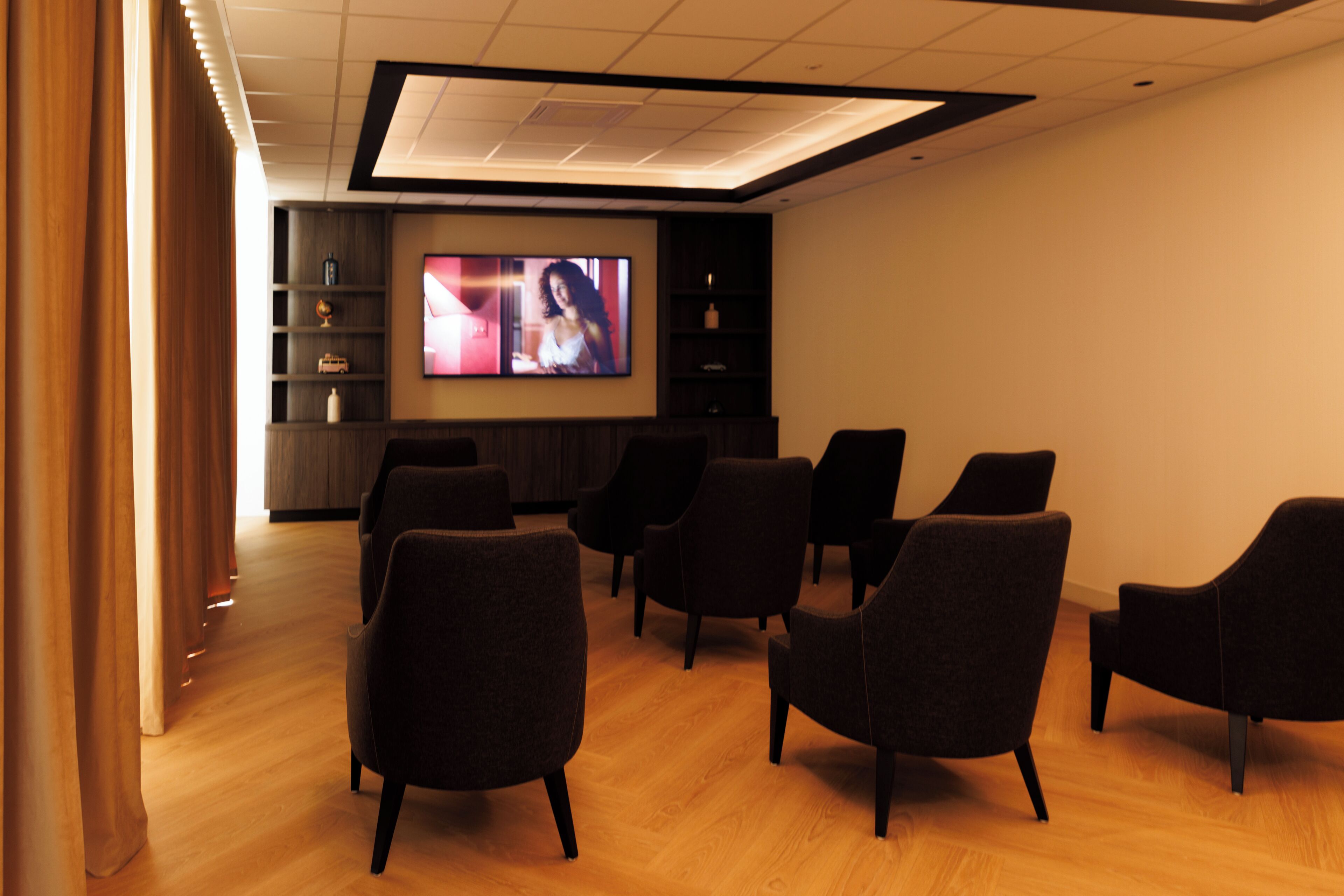 Quarto moderno com uma televisão de tela plana grande fixada na parede, várias cadeiras escuras dispostas em semicírculo e piso de madeira.