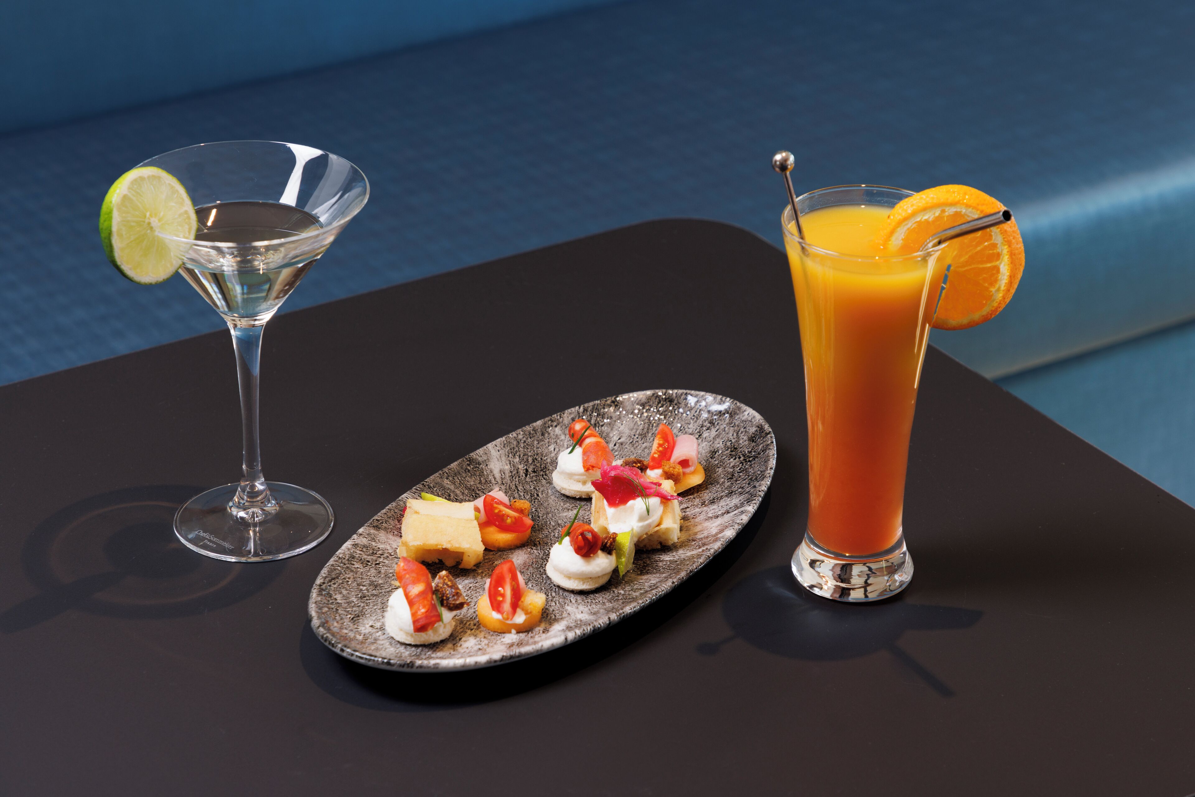 Martini-lasi ja korkea lasi appelsiinimehua pöydällä, jonka päällä on lautanen erilaisia alkupaloja.