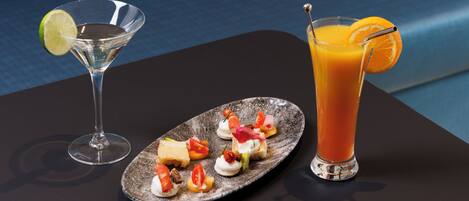 Una copa de martini y un vaso alto de jugo de naranja sobre una mesa, con un plato de aperitivos.