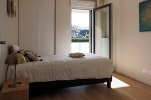 A bedroom with a bed, a nightstand, a lamp, a window with a view, and a painting on the wall.
