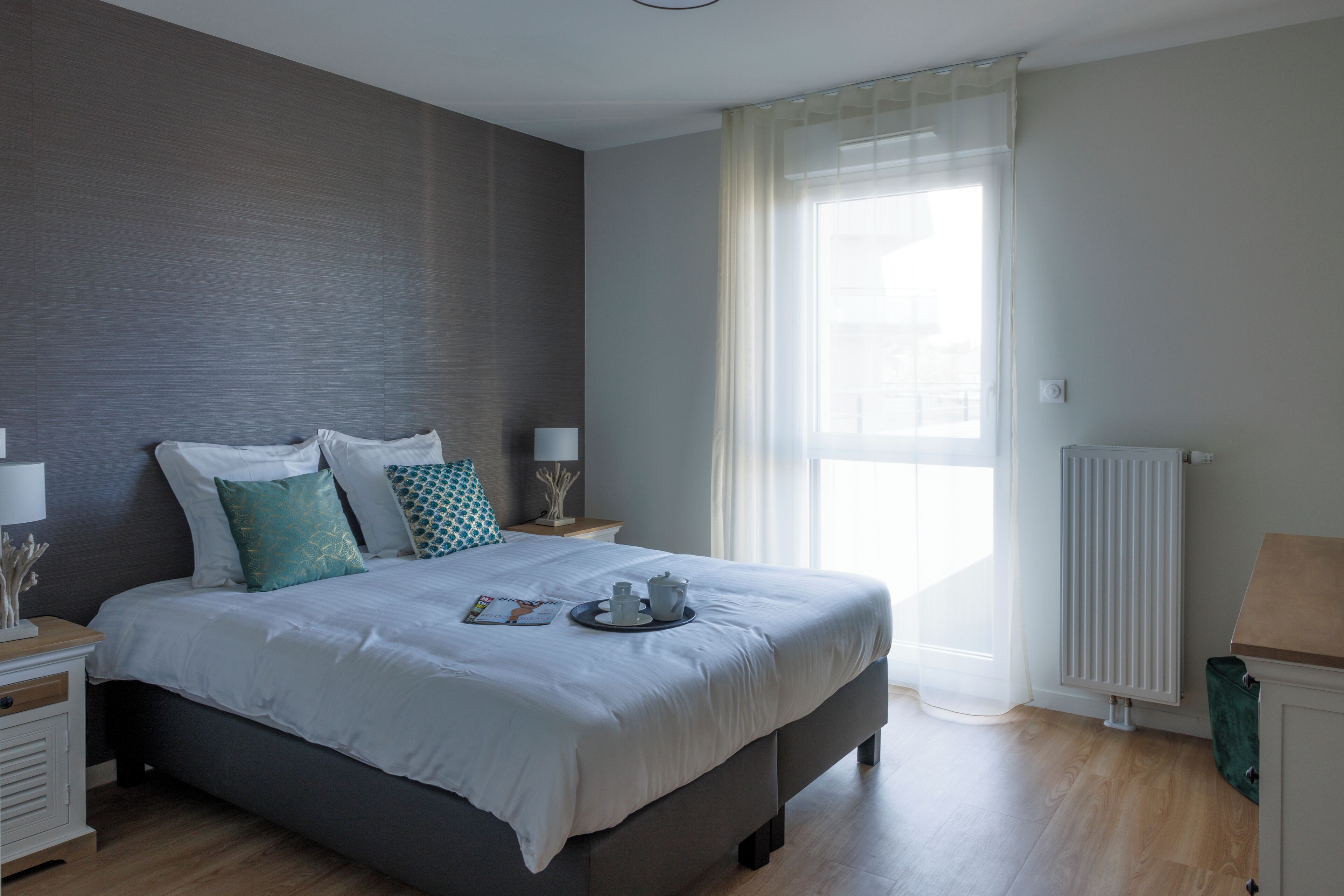 Un dormitorio moderno con una cama grande, mesitas de noche con lámparas, un radiador y una ventana con cortinas translúcidas.