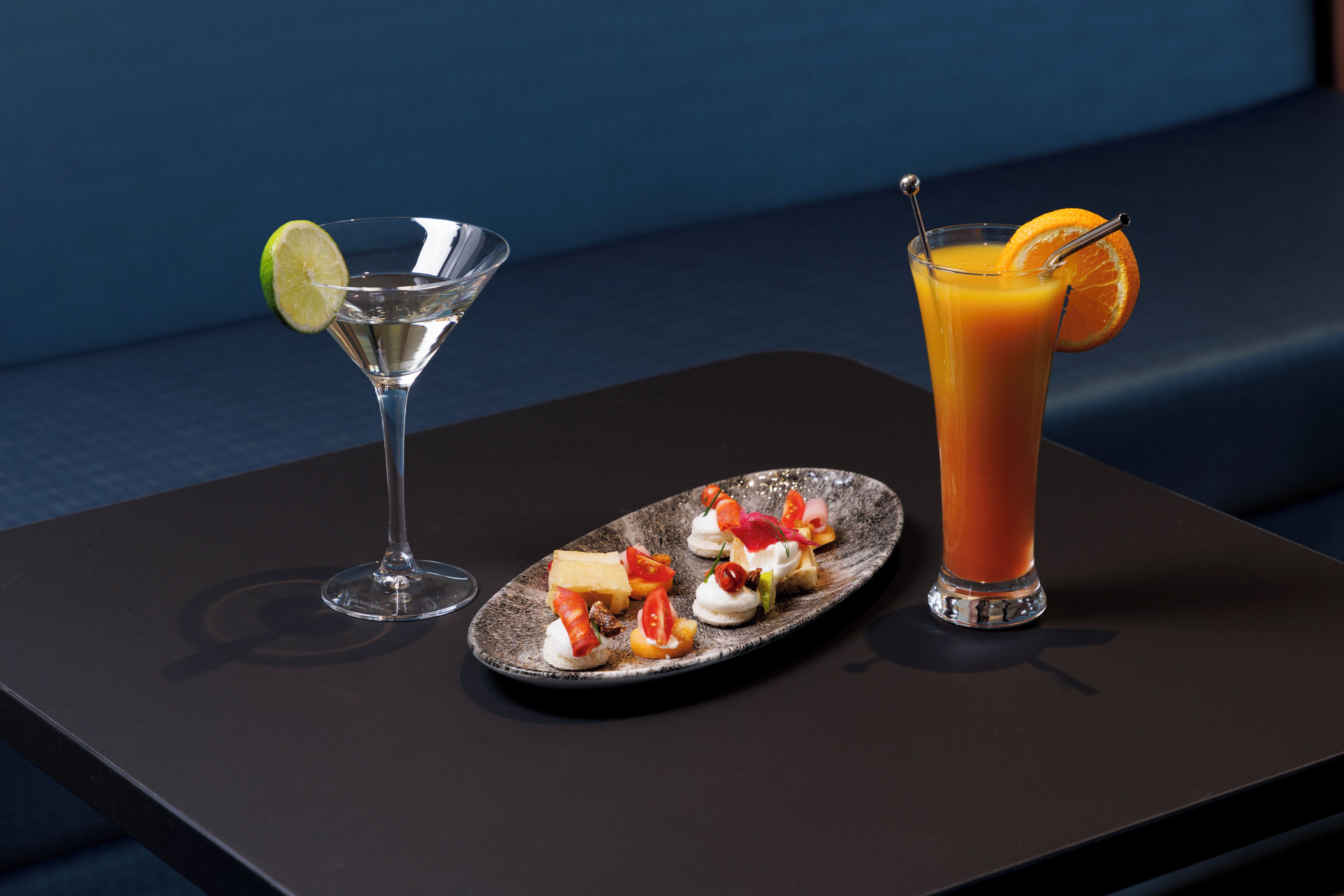 Una mesa con una copa de martini y un vaso alto de jugo de naranja, ambos con adornos, y un plato de aperitivos.