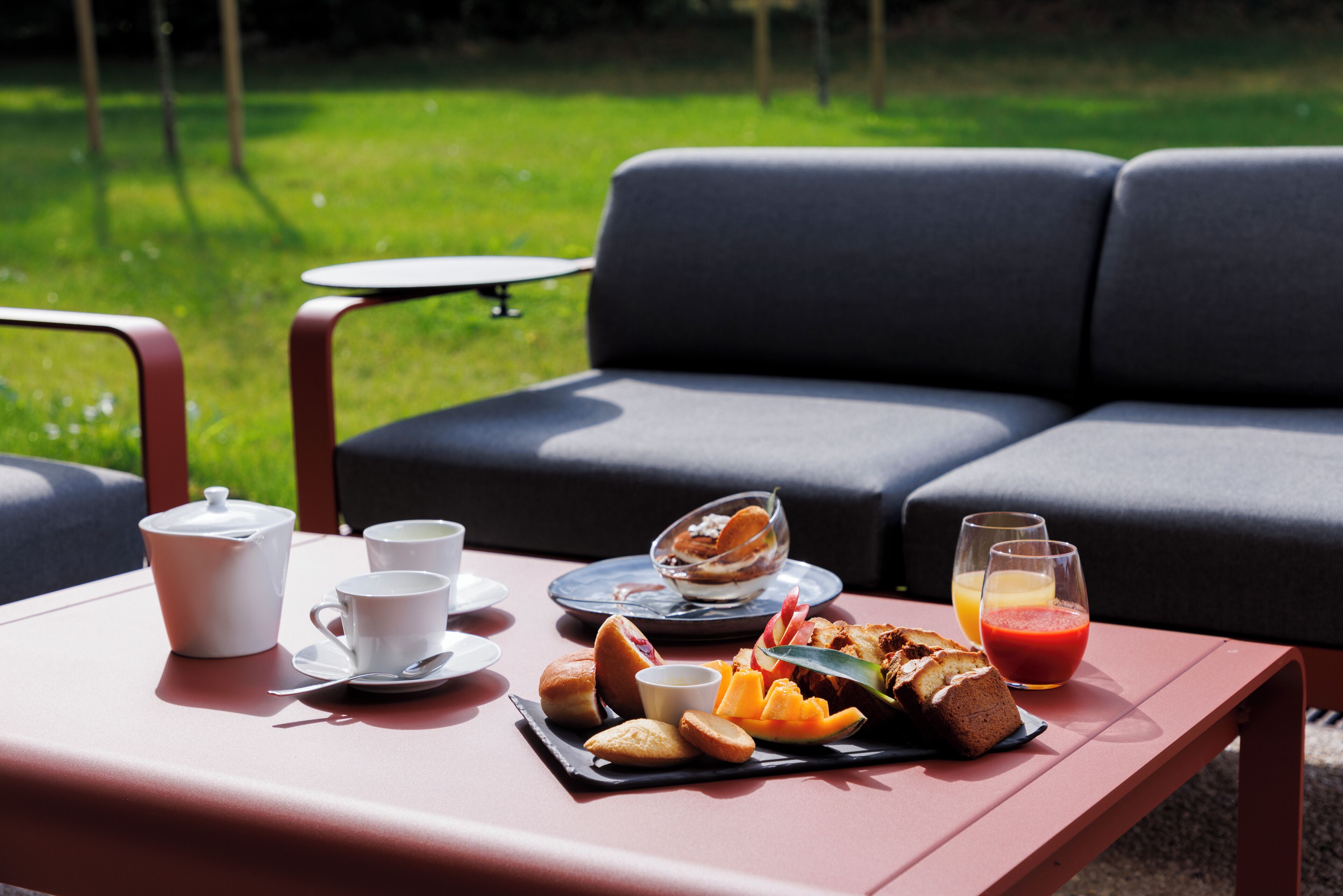 A table set with breakfast items, including bread, pastries, and juice, in an outdoor seating area with a green lawn and a sofa in the background.