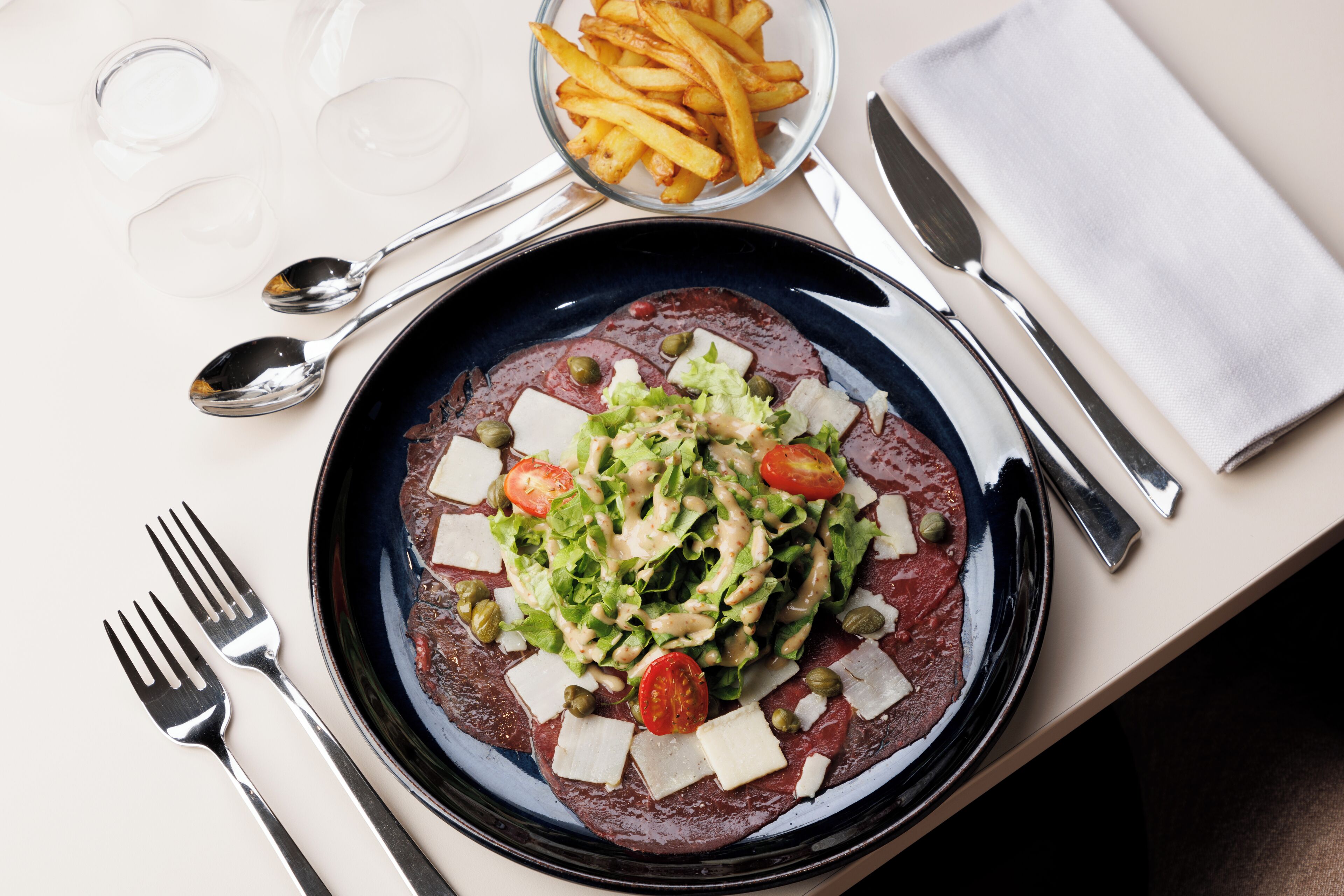 A plate of steak topped with lettuce, tomatoes, capers, and cheese, accompanied by a bowl of fries and cutlery on a table.