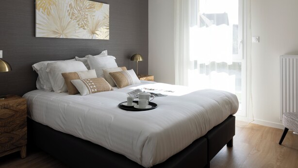 Een moderne slaapkamer met een groot bed, nachtlampjes, een kunstwerk aan de muur en een raam met dunne gordijnen.