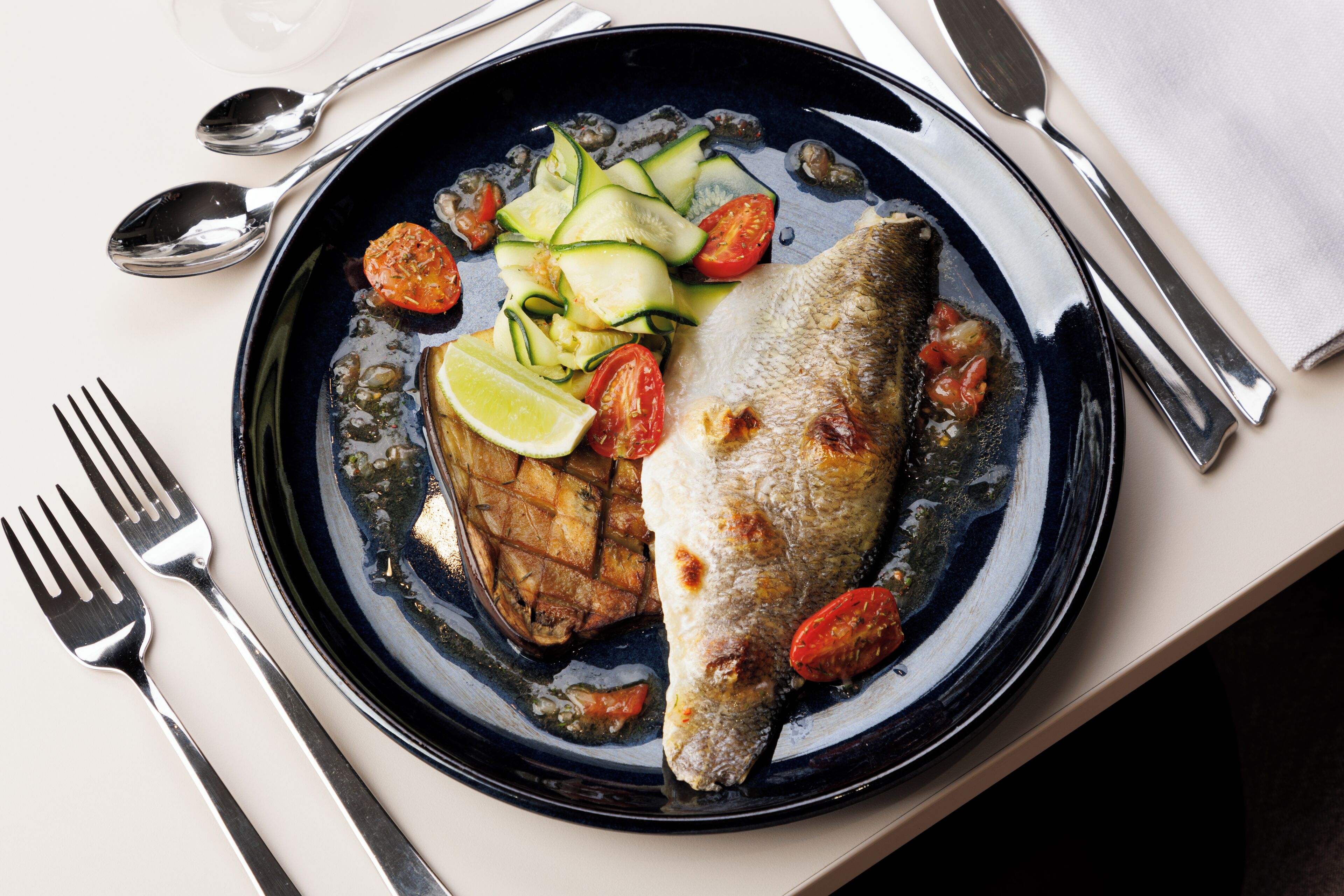 Un plato con pescado a la parrilla, tomates en rodajas y calabacín, acompañado de un gajo de lima.