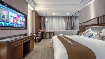 Habitación de hotel con una amplia televisión de pantalla plana montada en la pared, una cama con cabecera marrón, un escritorio con silla y una ventana con cortinas.
