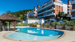 A modern hotel with a swimming pool, outdoor seating, and a thatched-roof structure. - Apto vista mar 4 dormitórios e piscina (Bombinhas)